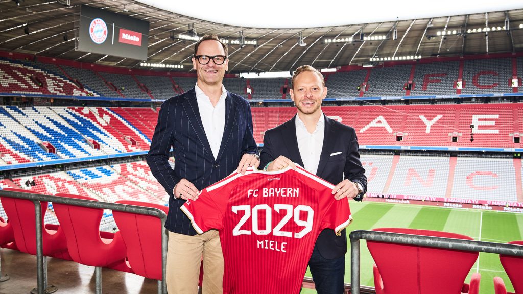 Rouven Kasper and Dr. Axel Kniehl hold up an FC Bayern shirt with “2029 Miele” at the Allianz Arena