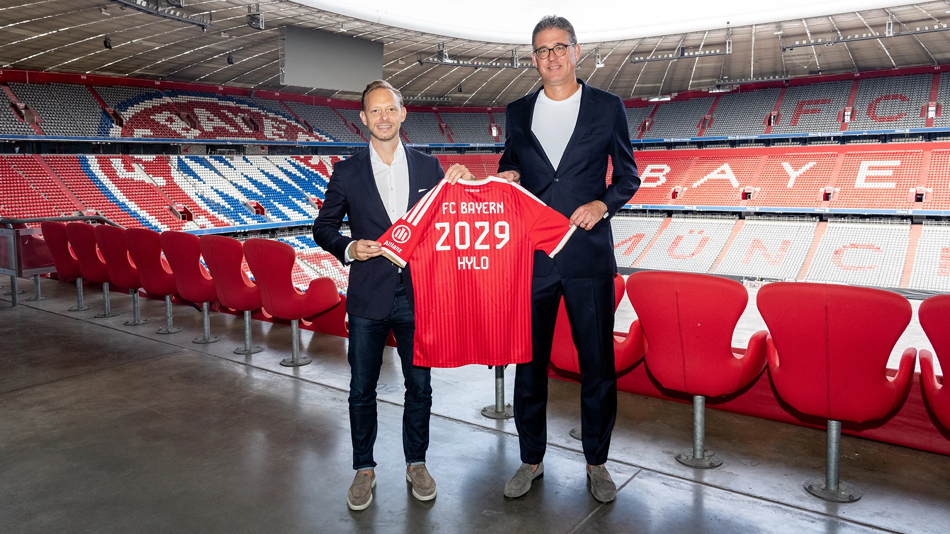 Rouven Kasper and Christian Krensel hold a red shirt with the words "FC Bayern 2029 HYLO" at the Allianz Arena