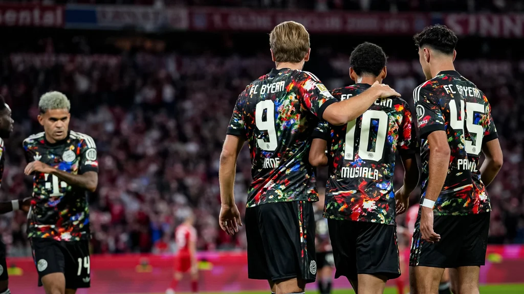 Harry Kane und Aleksandar Pavlović mit Jamal Musiala im Arm während des Bundesligaspiels zwischen dem FC Bayern und Union Berlin